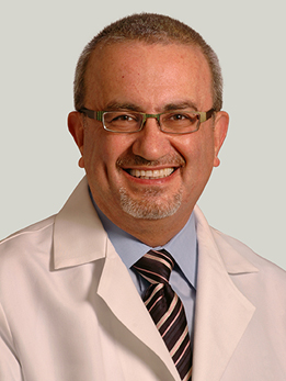 Issam Awad, MD, has short grey and brown hair and a brown goatee. He is wearing glasses, a blue dress shirt, a striped tie and a white lab coat. He is smiling broadly.