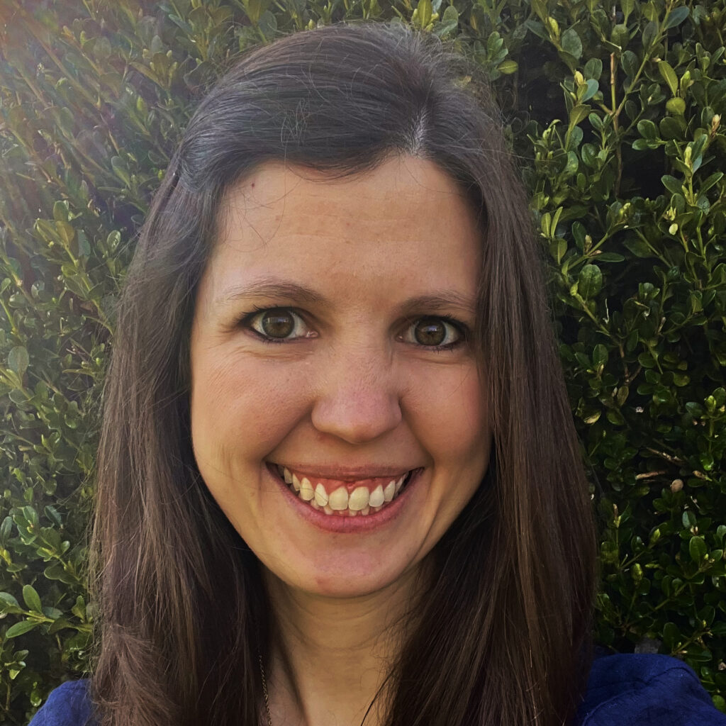 Photo of a woman smiling, she has long brown hair and brown eyes and is wearing a blue top.