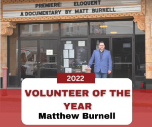 Photo of our Volunteer of the year, Matt Burnell. Matt is wearing a blue suit and standing in front of a marquee with the words: "PREMIERE ELOQUENT A DOCUMENTARY BY MATT BURNELL".