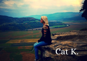 Woman with blonde hair sitting on the edge of a cliff with mountains and a sunrise in the background.