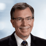 A headshot of smiling older white man with glasses wearing a suit