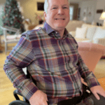 John Andres, an Alliance to Cure Board member. He has short white hair, is smiling, wearing a plaid shirt, and sitting in a wheelchair.