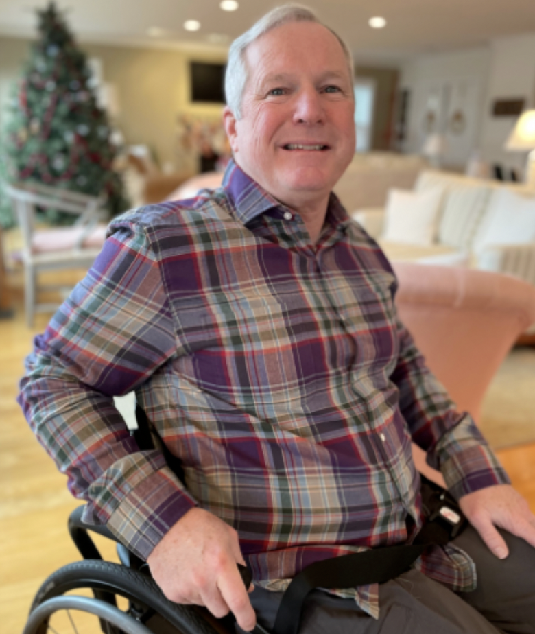 John Andres, an Alliance to Cure Board member. He has short white hair, is smiling, wearing a plaid shirt, and sitting in a wheelchair.