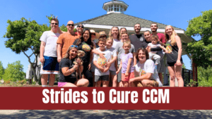 A large group of adults and children stand together smiling in front of a white gazebo on a sunny day, posing for a community photo. Some wear matching “Strides” event shirts, and one woman holds a small dog. A bold red banner across the bottom reads “Strides to Cure CCM,” highlighting the group’s participation in a walk to raise awareness and funds for cavernous malformation research.